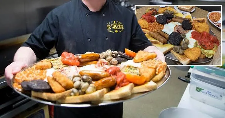 A chef in a black uniform with 'TO THIS CAFE' embroidered on the chest holds a large, overflowing tray of a traditional full…