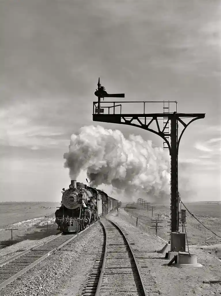 A black-and-white photograph shows a steam locomotive, numbered 1386, chugging down a railway track through a flat, open…