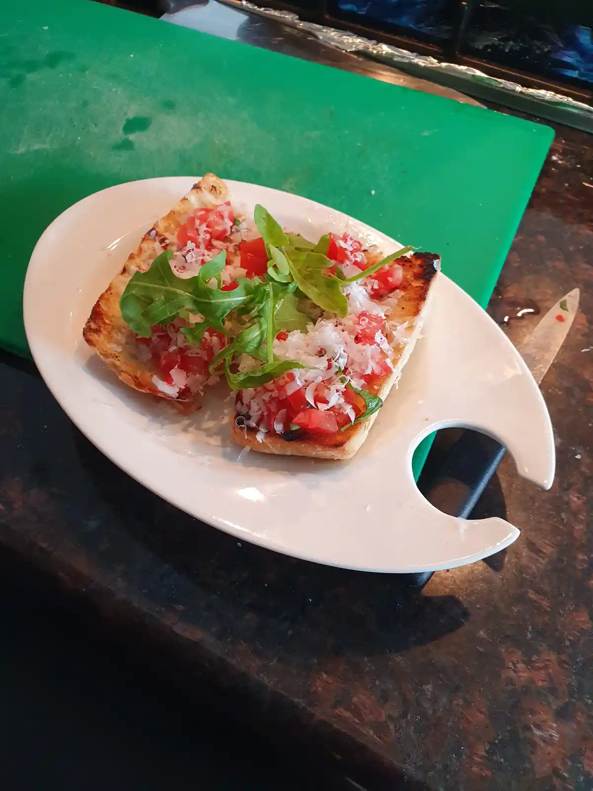 A white plate holds two slices of toasted bread topped with diced tomatoes, shredded cheese, and fresh arugula, sitting on a…
