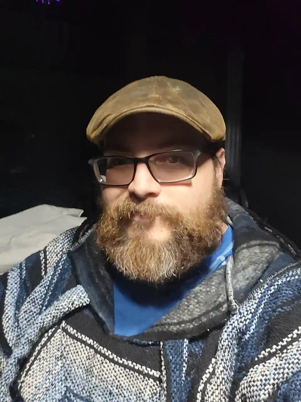 A man with a full beard, glasses, and a brown flat cap looks directly at the camera with a neutral expression while wearing…