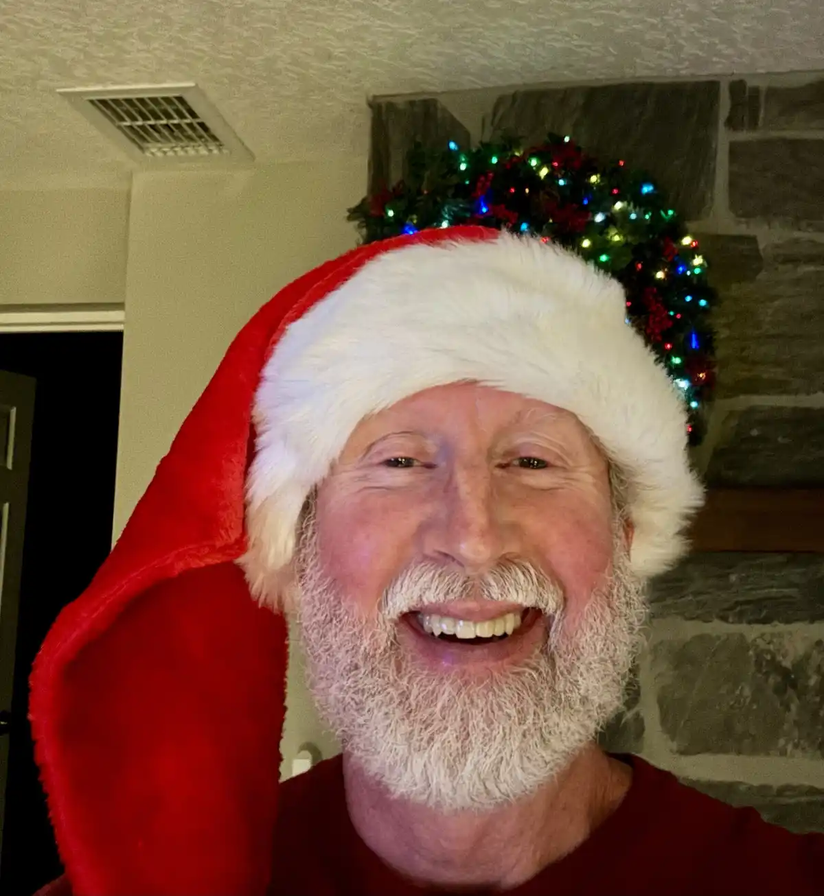 A cheerful older man with a white beard wears a red and white Santa hat while smiling broadly at the camera. Behind him, a…