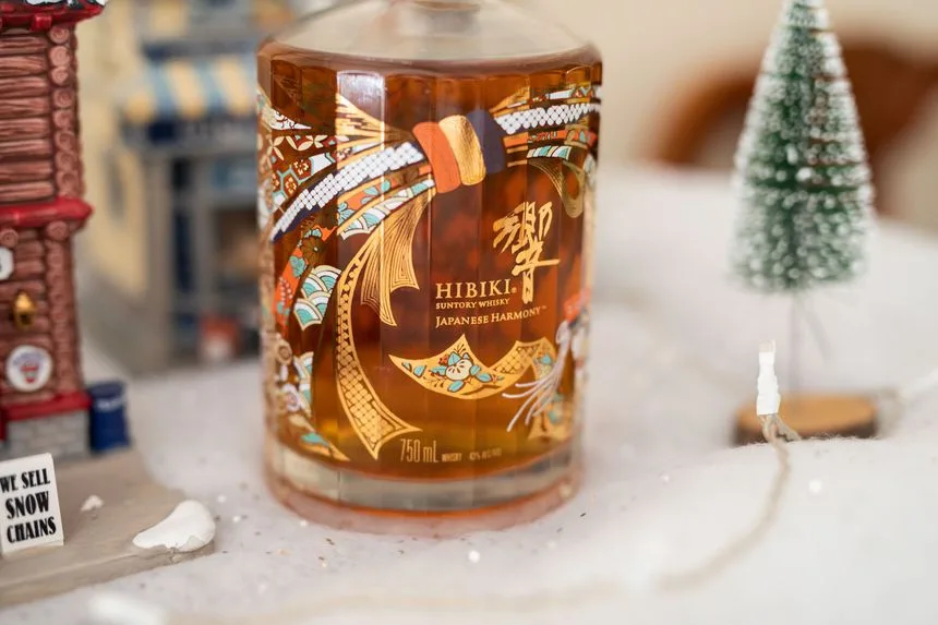 A bottle of Hibiki Suntory Whisky Japanese Harmony sits in a snowy diorama with miniature buildings and a small Christmas…