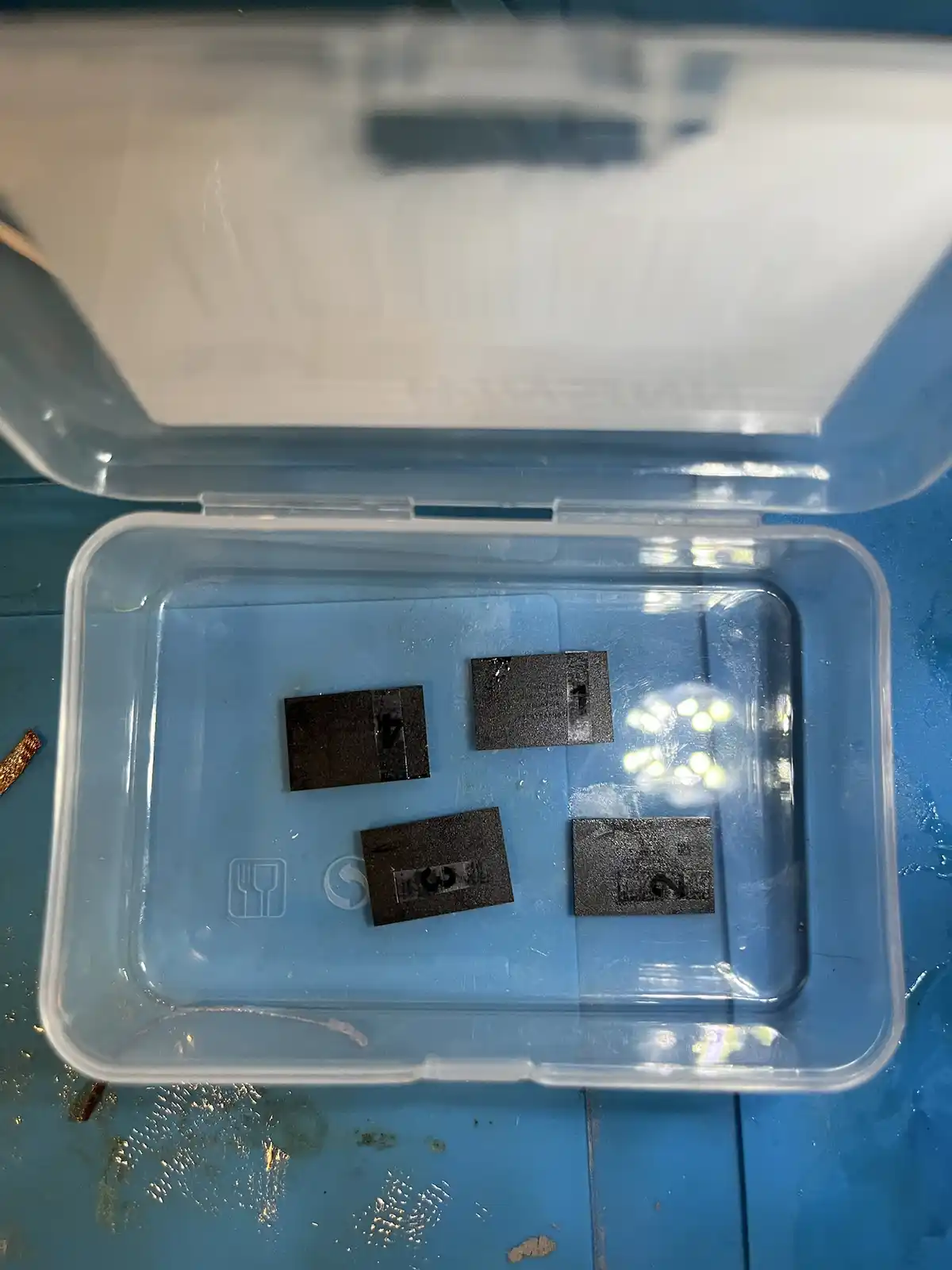 An overhead shot shows four small, dark rectangular metal pieces inside a clear plastic container, each labeled with a…