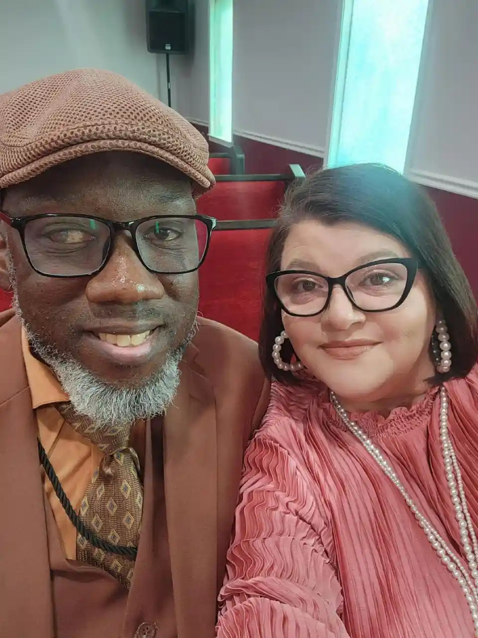 A man and a woman are posing together for a selfie in what appears to be a theater. The man wears a brown flat cap, glasses…