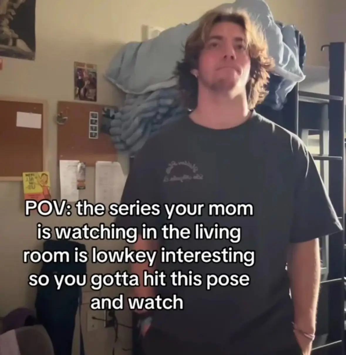 A young man with shoulder-length wavy hair and a black t-shirt stands in a dorm room with a messy bunk bed behind him. He…