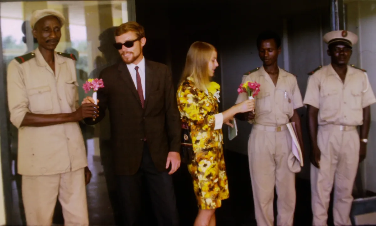 A vintage photo shows a man in a suit and sunglasses smiling while accepting a small bouquet of pink and yellow flowers from…