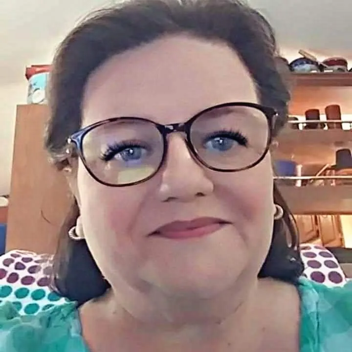A woman with dark-rimmed glasses and a subtle, knowing smile looks directly at the camera. She wears a green top with a…