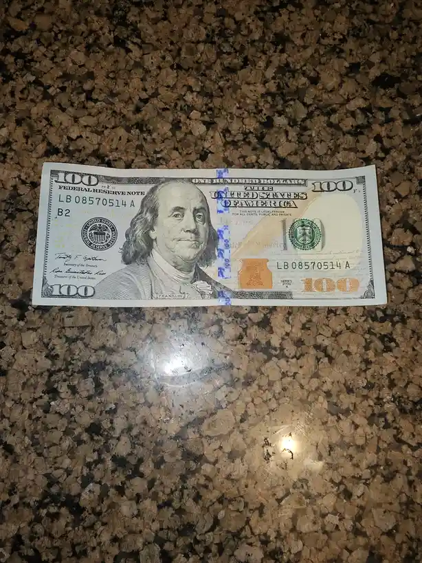 A U.S. $100 bill is laid flat on a speckled brown countertop, with a large, orange, handwritten '1' drawn over the right…