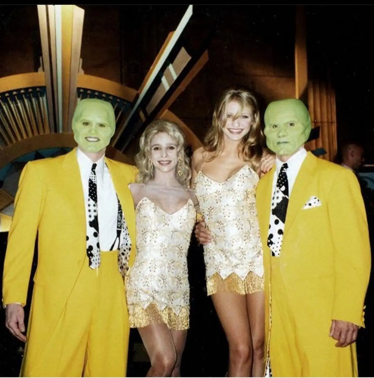 Two men in identical green The Mask masks and bright yellow suits flank two smiling women in gold-patterned dresses at what…