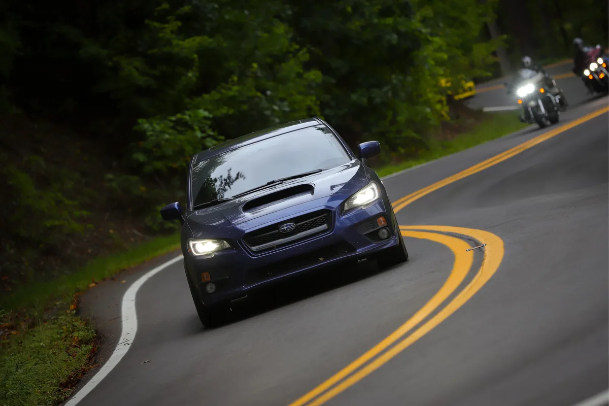 A blue Subaru WRX STI is driving on a curving road with trees on the side, and two motorcycles are visible behind it. The…
