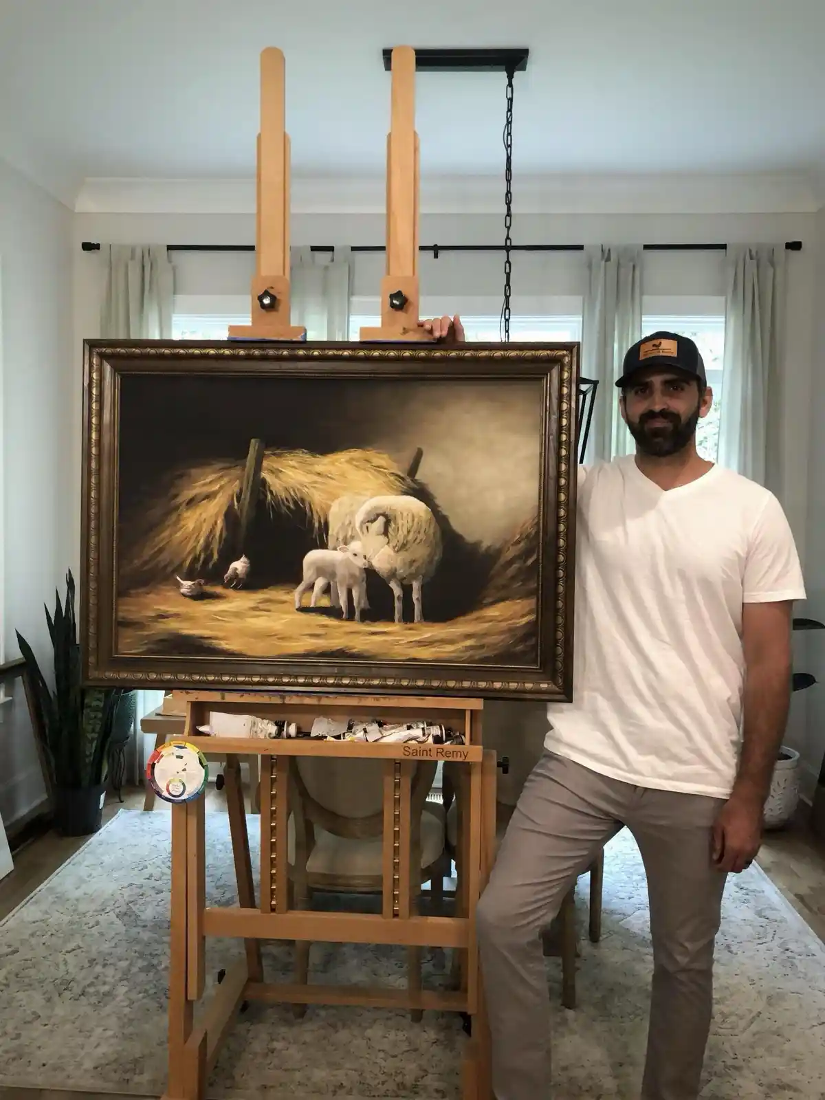 A man in a white t-shirt and baseball cap stands beside a large, ornate wooden easel holding a framed painting of sheep and…