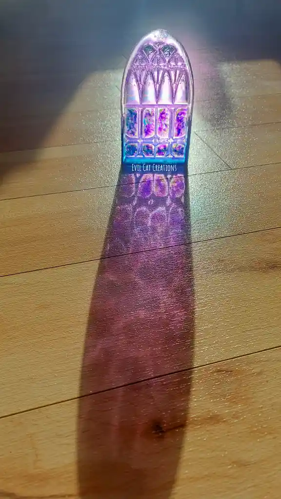 A small, illuminated, arched stained-glass window-shaped object sits on a wooden floor, casting a long, colorful, shimmering…