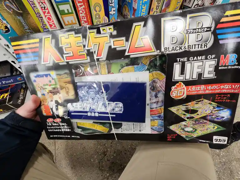 A person holds a Japanese board game box for '人生ゲーム BB' (The Game of Life) with 'BLACK & BITTER' branding, which is a…