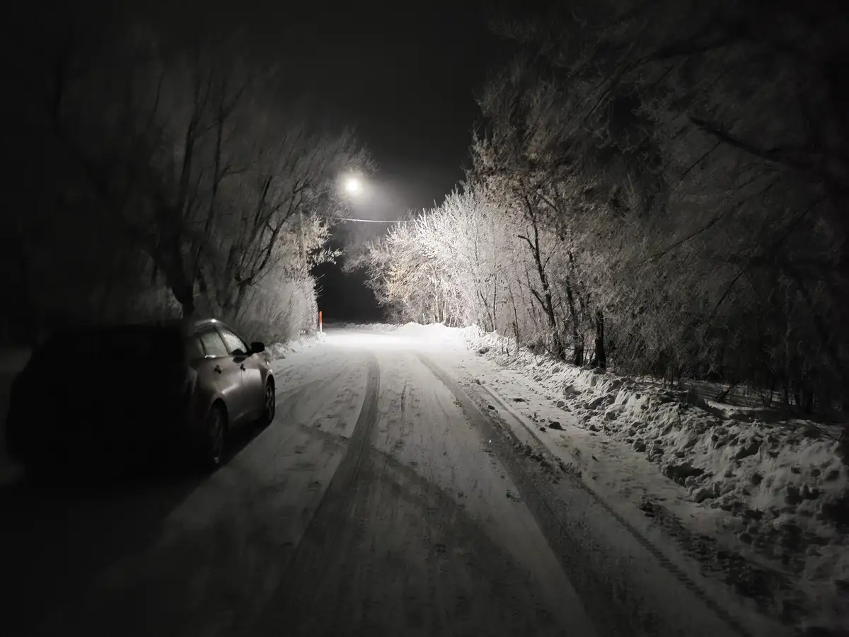 A car is parked on a snow-covered road at night, illuminated by a streetlight, with frost-covered trees lining the path. The…