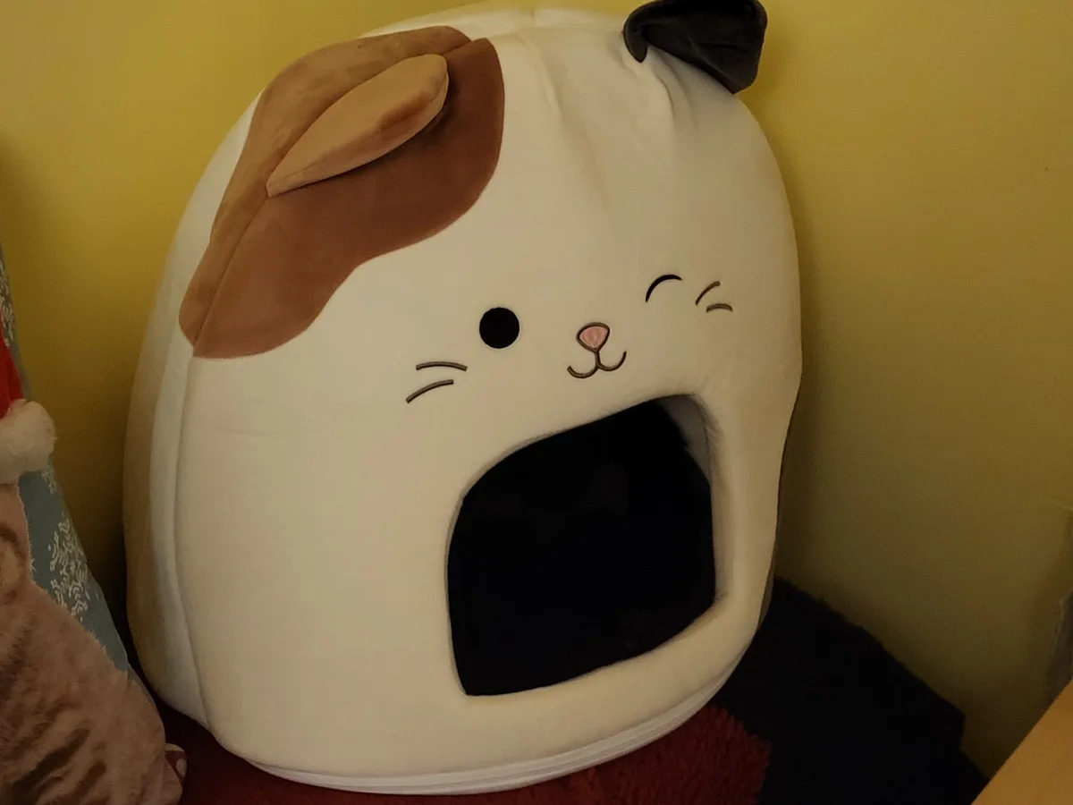 A plush, cat-shaped pet bed with a white body, brown ear, and black ear, featuring a smiling face with closed eyes and a…