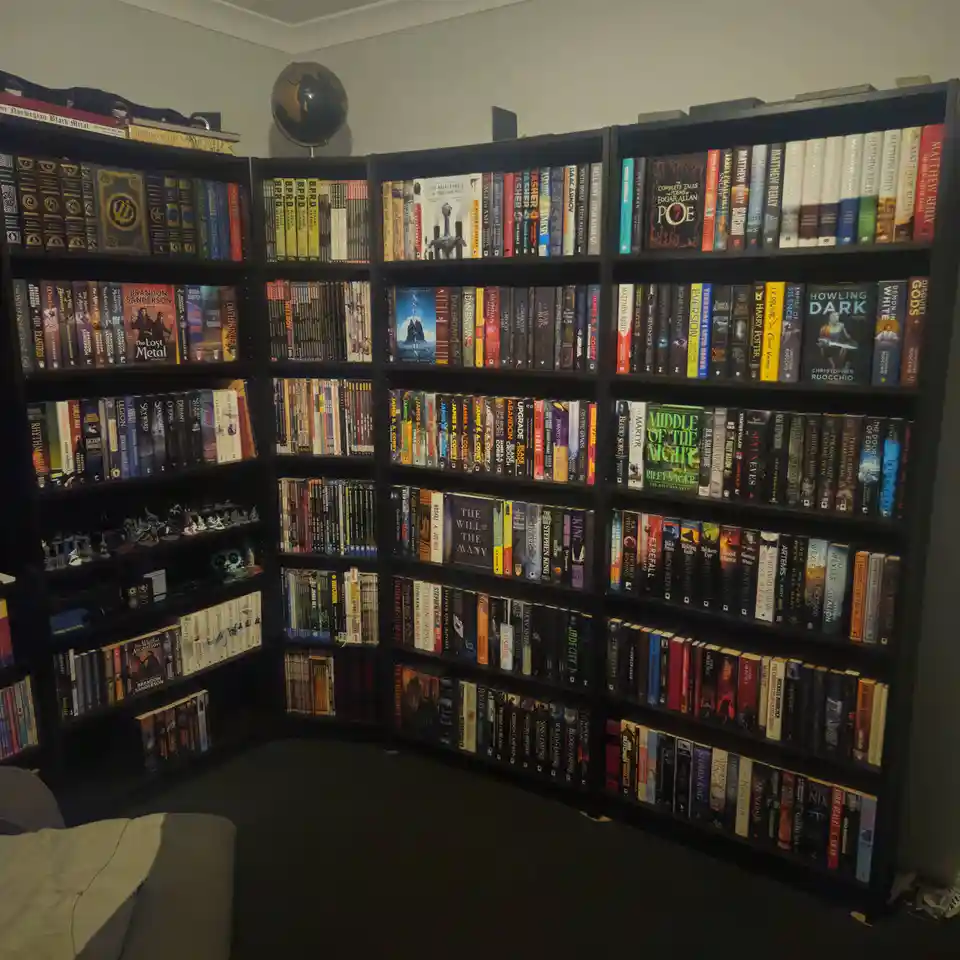 A corner of a room filled with tall, black bookshelves packed with hundreds of books, mostly fantasy and sci-fi, with a…