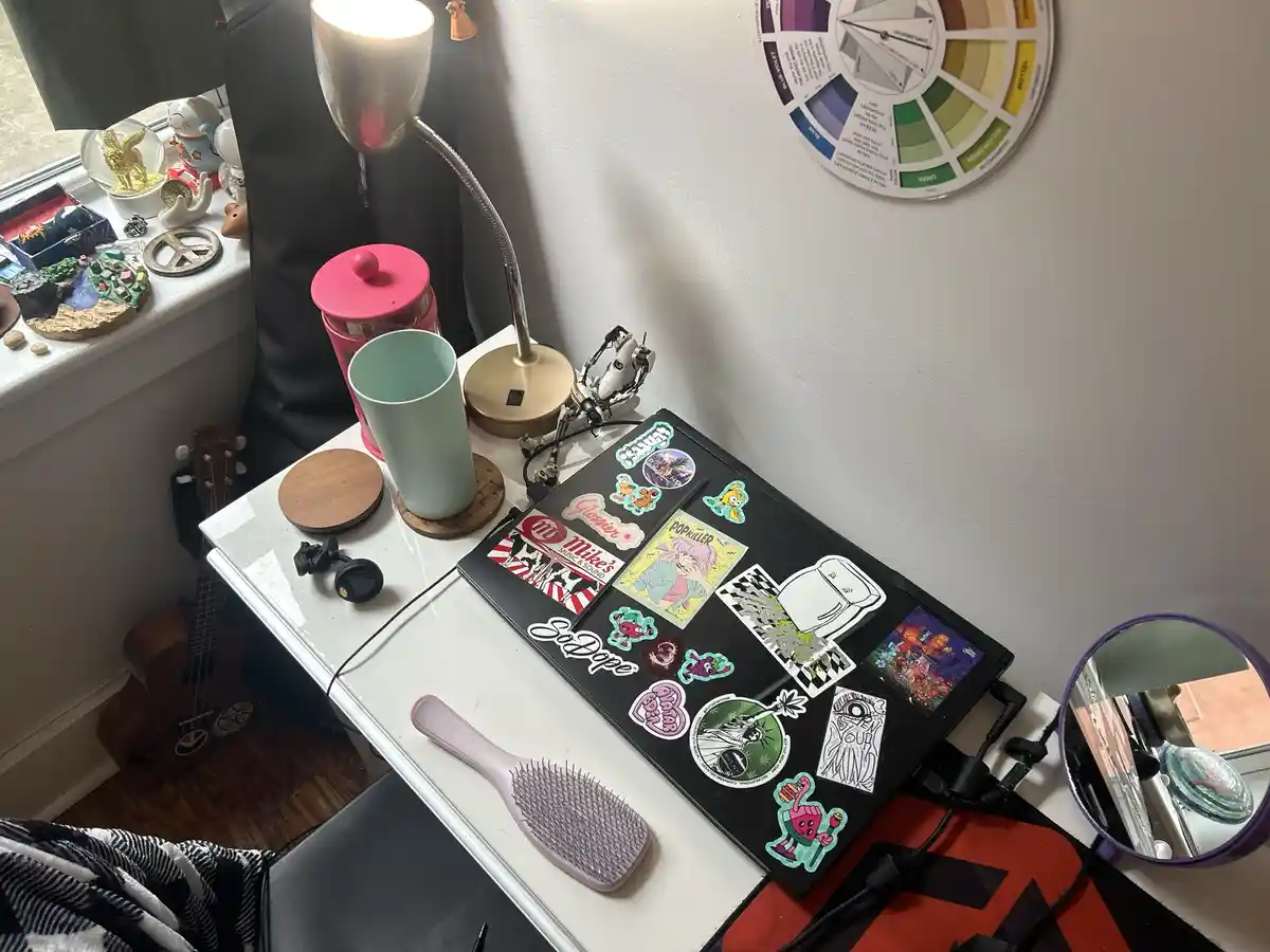 A white desk is cluttered with a laptop covered in colorful stickers, a pink mug, a lamp, a ukulele, and a hairbrush. A…