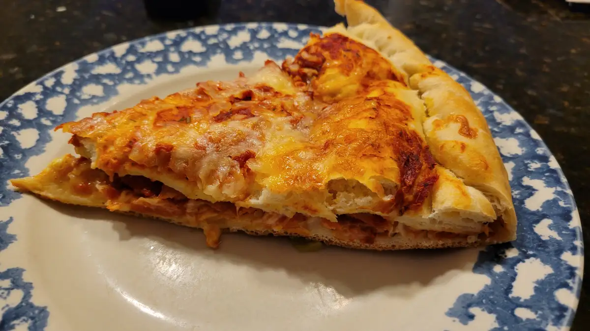A close-up photo shows a single slice of pizza on a white plate with a blue speckled rim, resting on a dark countertop. The…