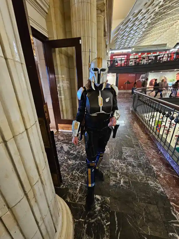 A person in a detailed Mandalorian cosplay, featuring a helmet with gold and white accents, black armor with gold trim, and…