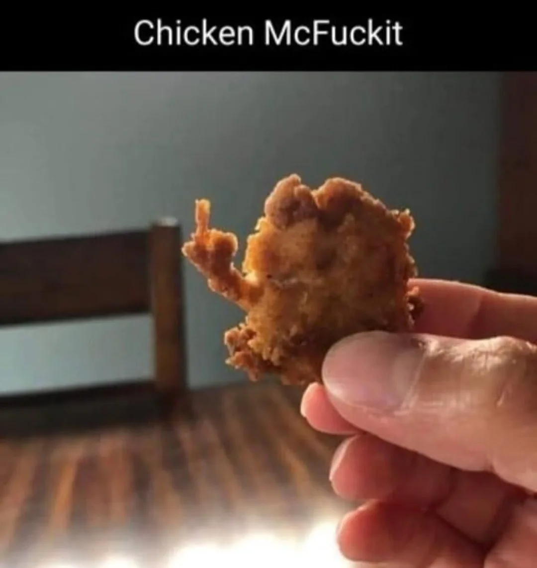 A hand holds a piece of fried chicken that has an irregular, lumpy shape with a protruding piece resembling a finger…