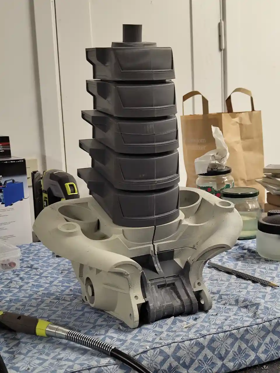 A 3d printed object resembling a tower of stacked plastic drawers sits on a blue patterned surface, surrounded by tools and…