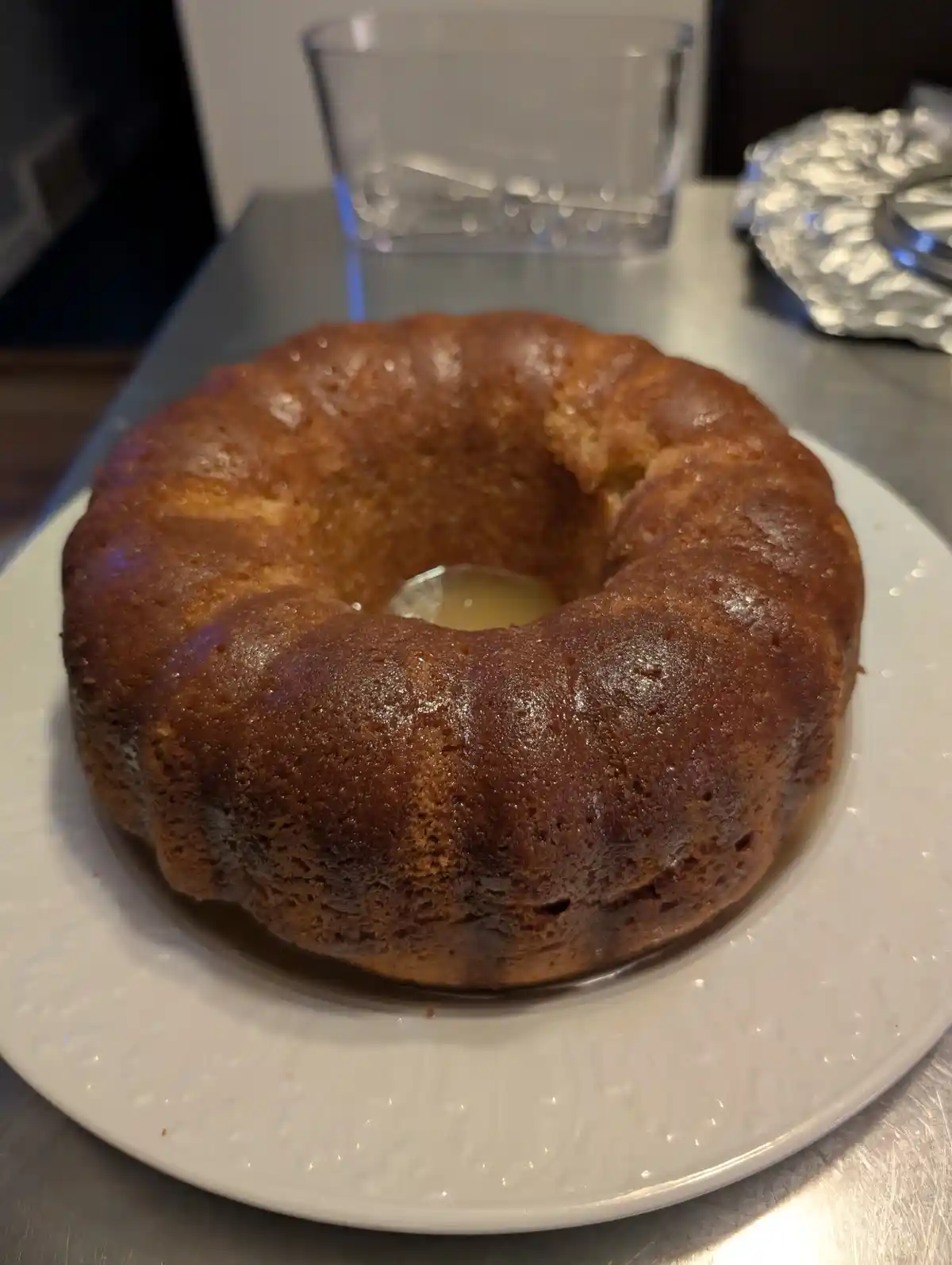 A golden-brown bundt cake sits on a white plate, with a pool of syrup in its center, placed on a stainless steel countertop…