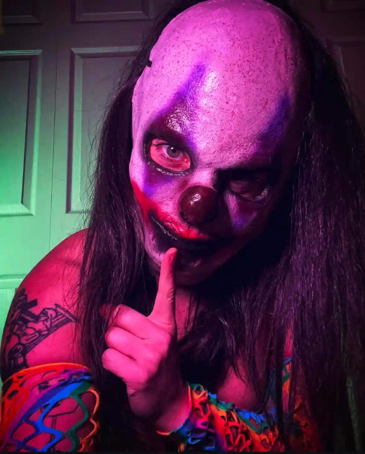 A person with long dark hair wears a grotesque, textured clown mask with purple and red lighting, a red nose, and darkened…