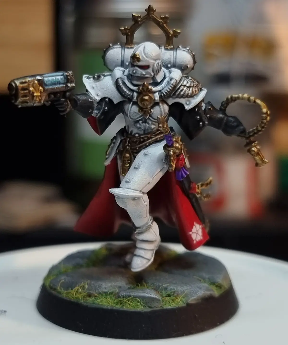 A painted miniature of a Space Marine in ornate silver and gold armor, holding a bolter in one hand and a chain weapon in…