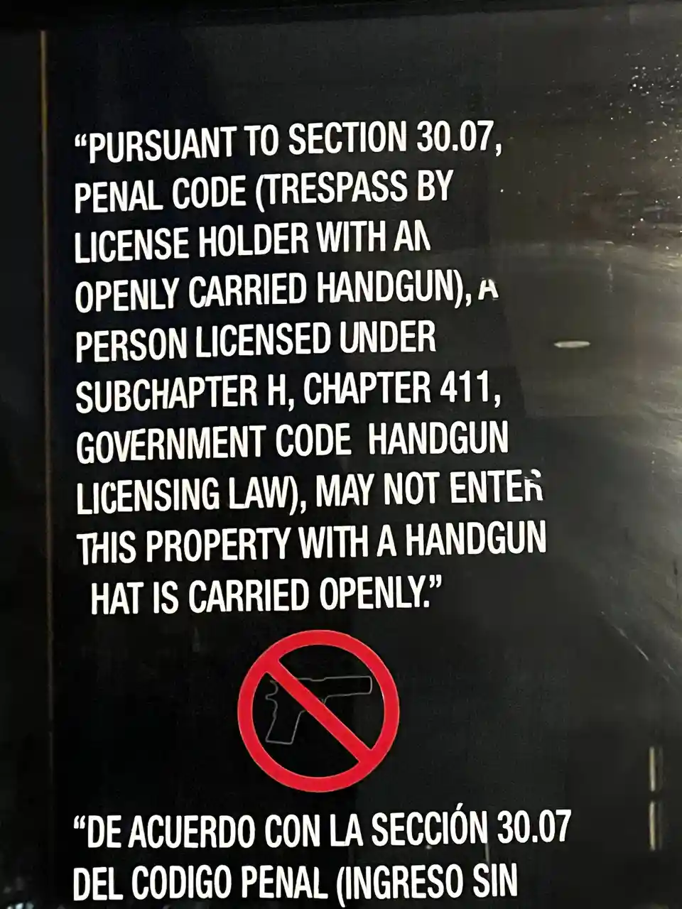 A black sign with white text and a red 'no guns' symbol reads a legal notice prohibiting handgun carry on property, but the…