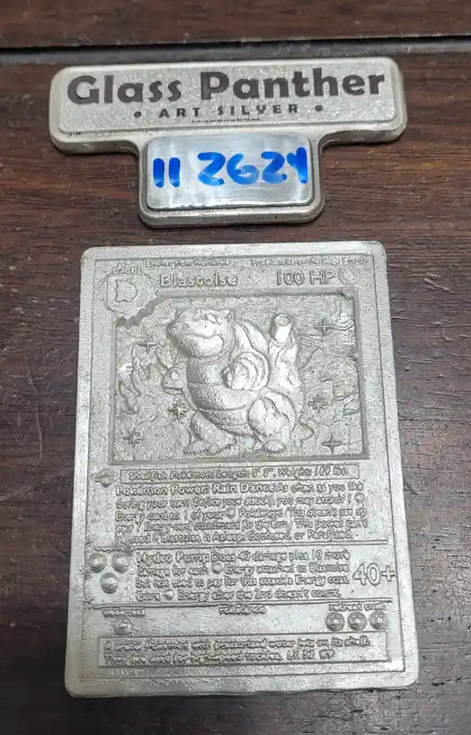 A silver-colored, embossed physical replica of a Blastoise Pokémon trading card is displayed on a wooden surface, next to a…