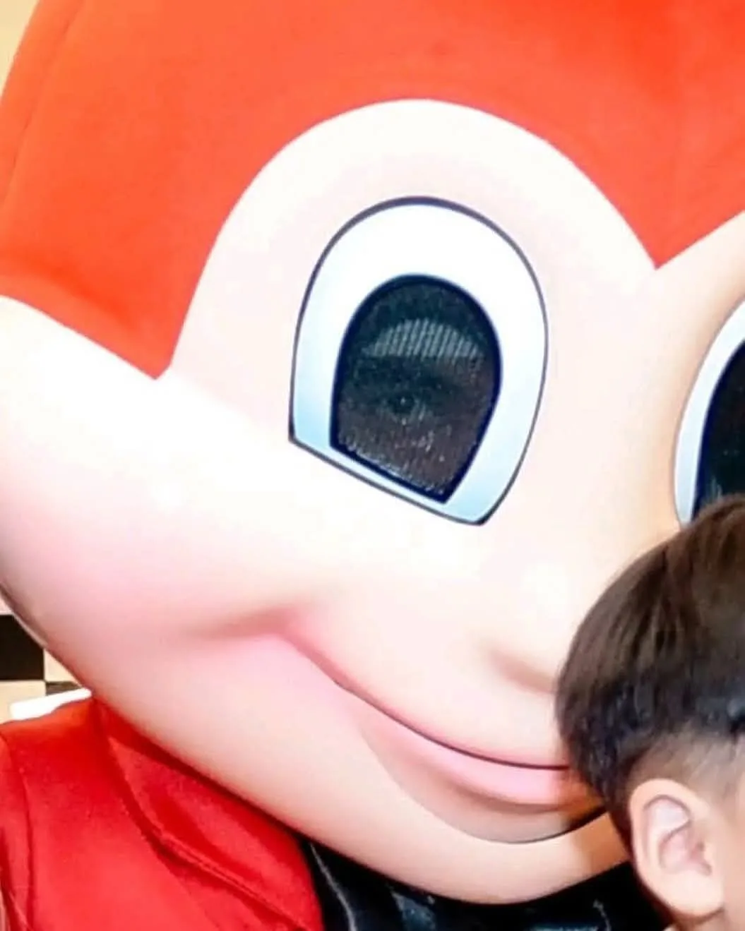 A close-up photo shows the face of a large, red, cartoonish mascot with big, round eyes and a smiling mouth. In the bottom…