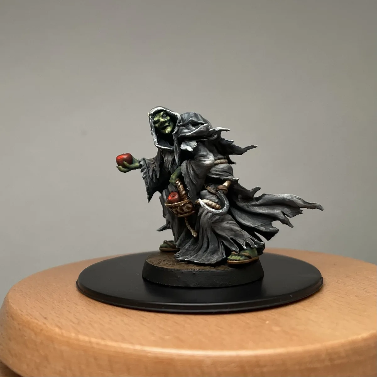 A painted miniature figurine of a green-skinned, hooded demon-like creature holding a red apple in one hand and a basket of…