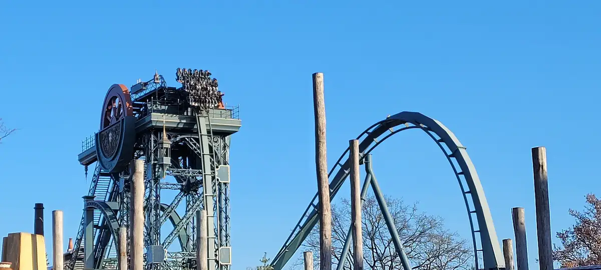 A roller coaster with a tall, intricate tower structure and a large loop against a clear blue sky. Riders are visible at the…