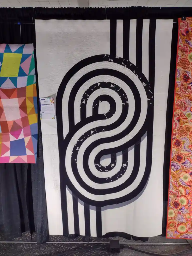 A large black and white quilt featuring a stylized, swirling 'S' shape with concentric loops and vertical stripes, displayed…