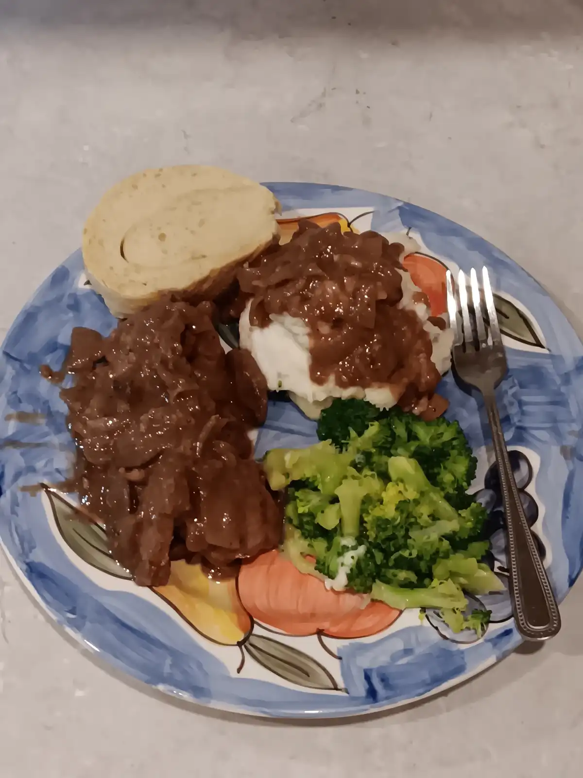 A plate of food featuring beef stroganoff with a thick brown sauce, mashed potatoes, steamed broccoli, and a roll, all on a…