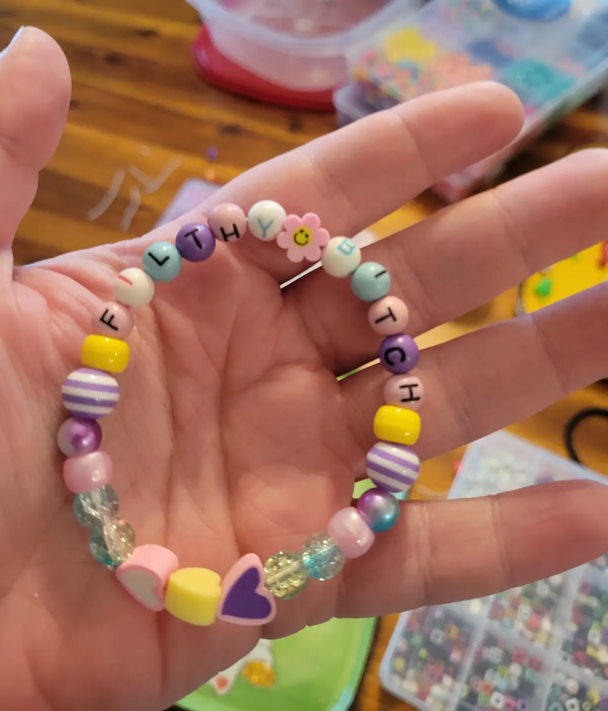 A hand holds a colorful beaded bracelet made of various pastel beads, some with letters spelling 'HEALTHY' and 'BITCH', a…