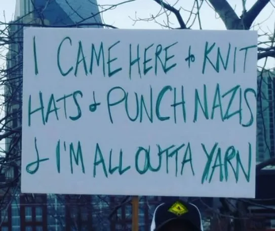 A white cardboard sign with handwritten teal text reads 'I CAME HERE to KNIT HATS & PUNCH NAZIS & I'M ALL OUTTA YARN'. The…