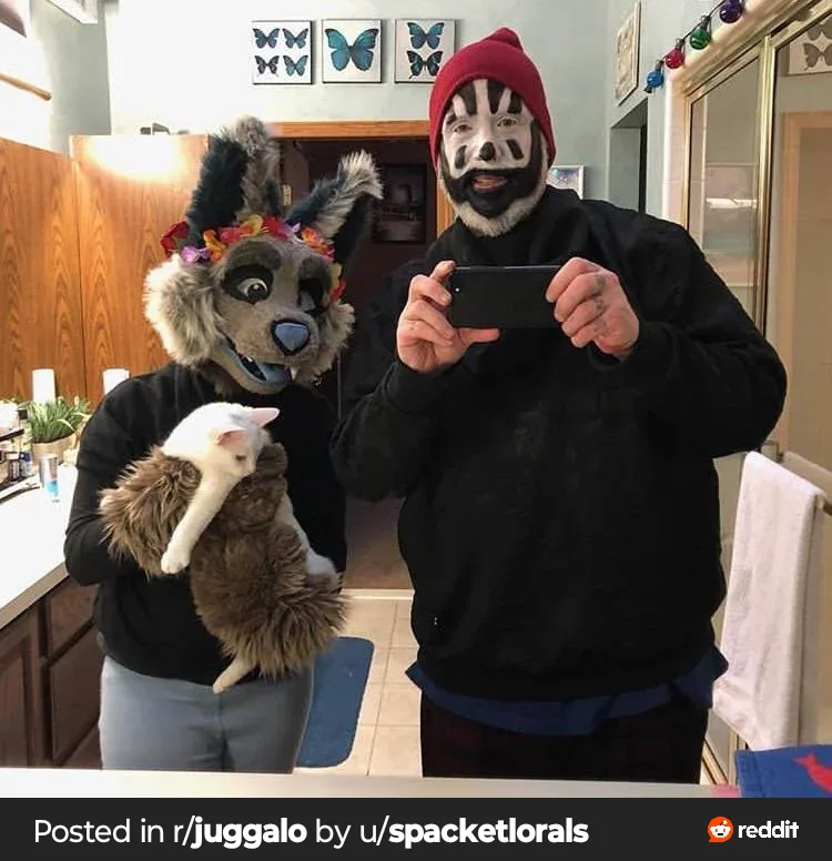 Two people in a bathroom mirror selfie: one wears a furry wolf-like costume holding a white cat, the other wears a red…