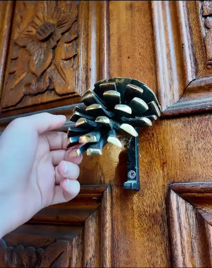 A hand holds a pinecone that has been fashioned into a door knocker, mounted on a dark wooden door with carved floral…