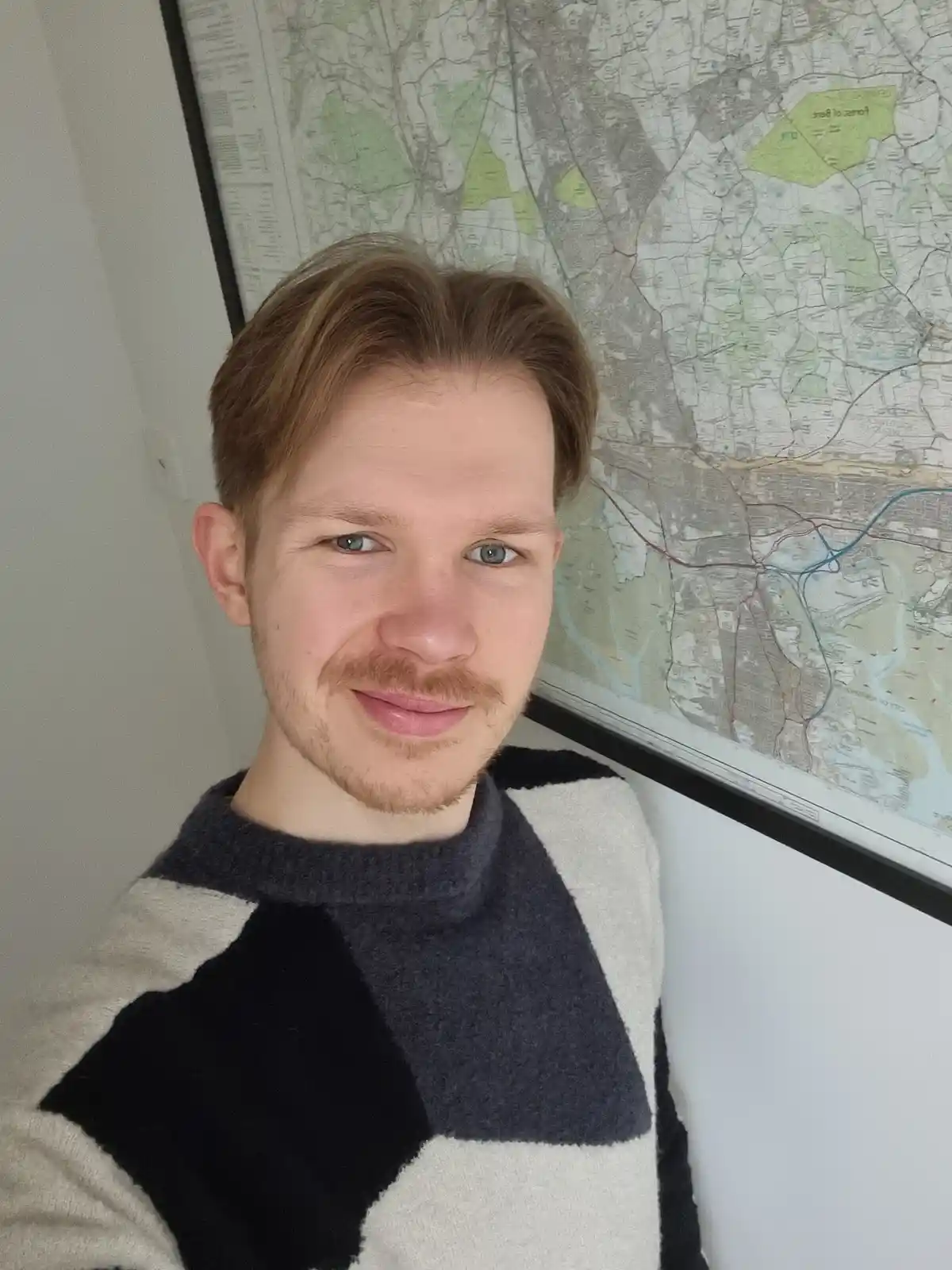 A young man with light brown hair and a mustache smiles at the camera while standing in front of a large framed map on a…