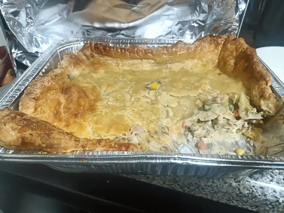 A rectangular aluminum baking pan holds a golden-brown, flaky-crust pie with a creamy filling visible in a partially eaten…