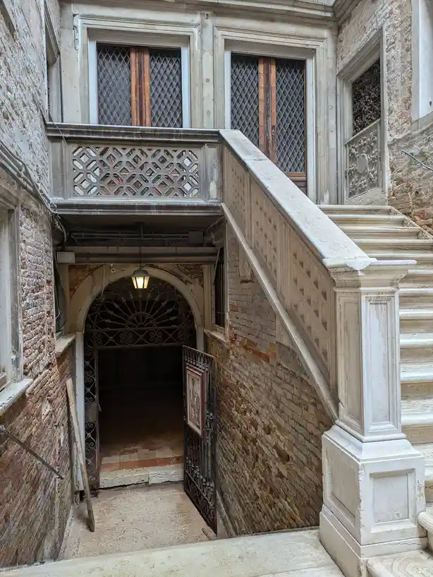 An image of a grand, ornate stone staircase leading up to a balcony with diamond-paned windows, next to a lower arched…