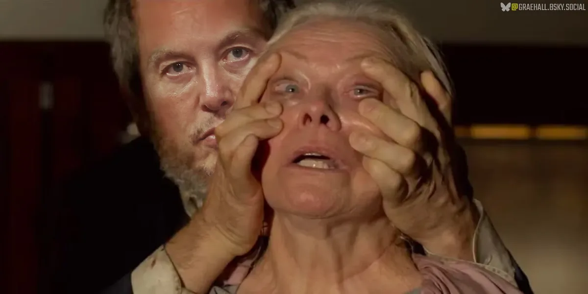 A man with a beard holds an older woman's face with both hands, pulling her eyelids open as she looks upward with a…