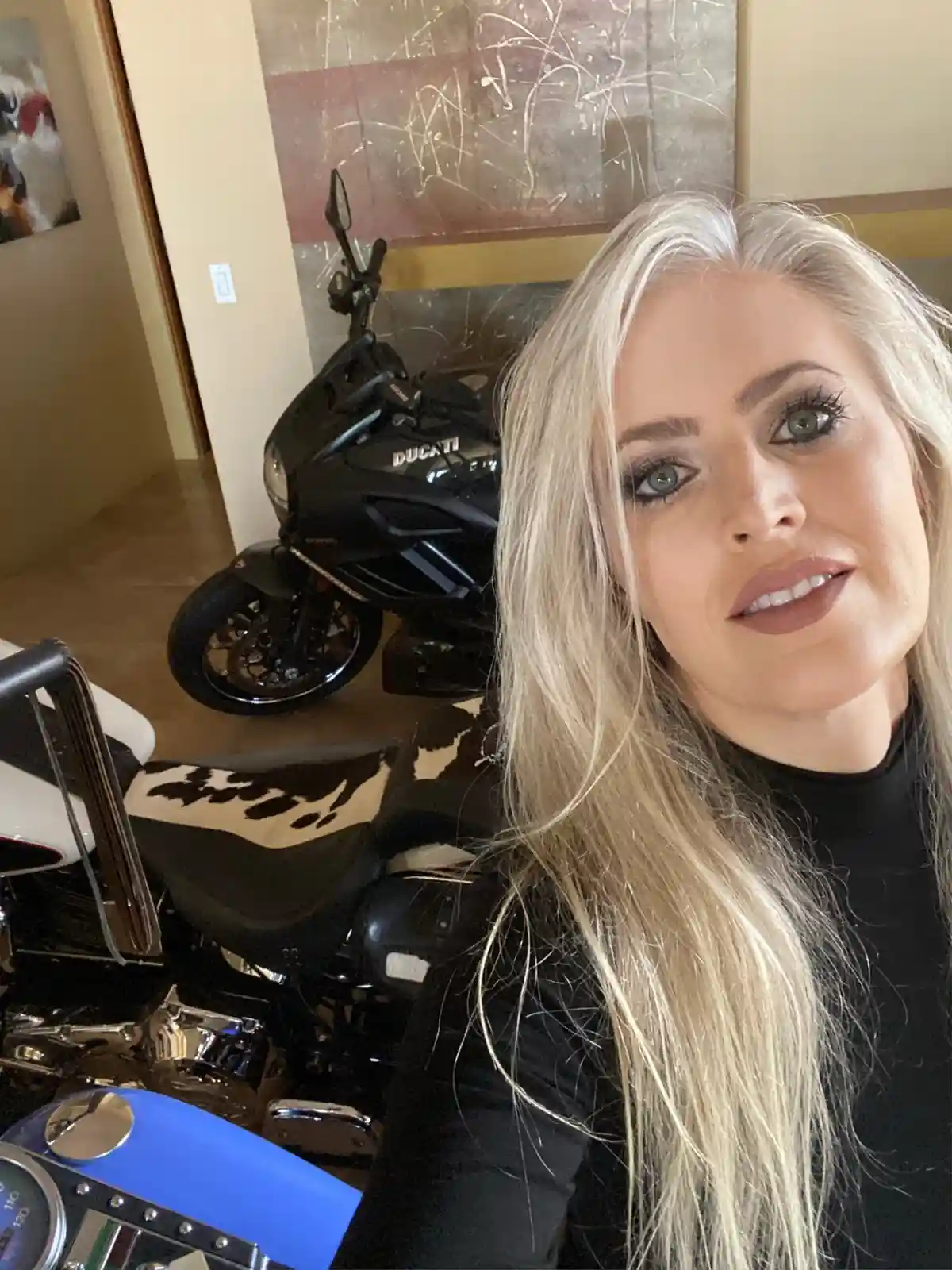 A woman with long blonde hair and a black top poses for a selfie indoors, with two motorcycles visible behind her: a black…
