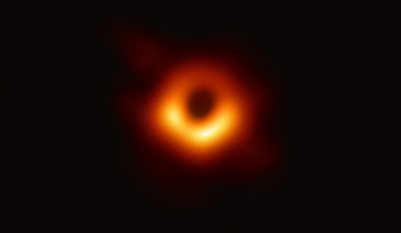 The image shows a dark, circular void surrounded by a bright orange ring of light against a black background, resembling the…