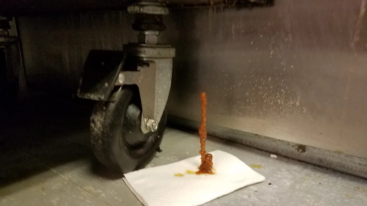 A close-up photo shows a thick, orange-brown sauce dripping vertically from a metal caster wheel under a kitchen appliance…