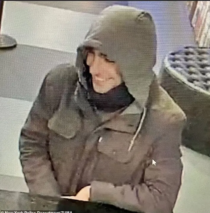 A grainy security camera image shows a person in a dark hooded jacket and scarf, smiling while seated at a counter. The…
