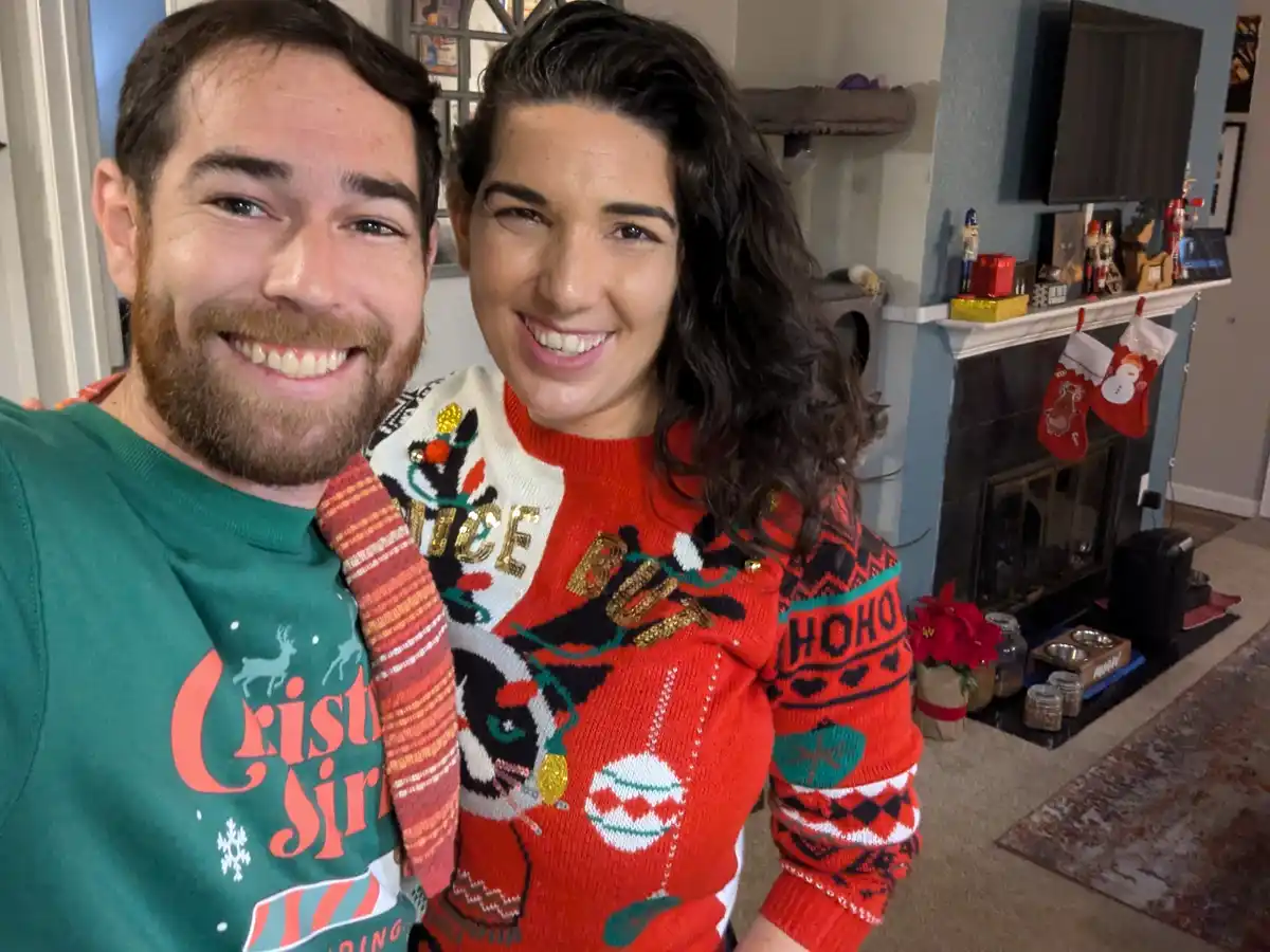 A smiling man and woman pose for a selfie in a living room decorated for Christmas, both wearing festive sweaters. The man…