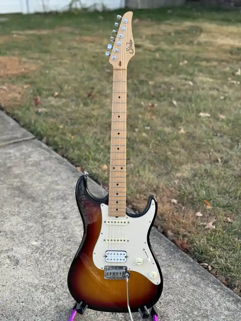 A sunburst-finish electric guitar, likely a Squier Stratocaster, stands upright on a concrete sidewalk next to grass. The…