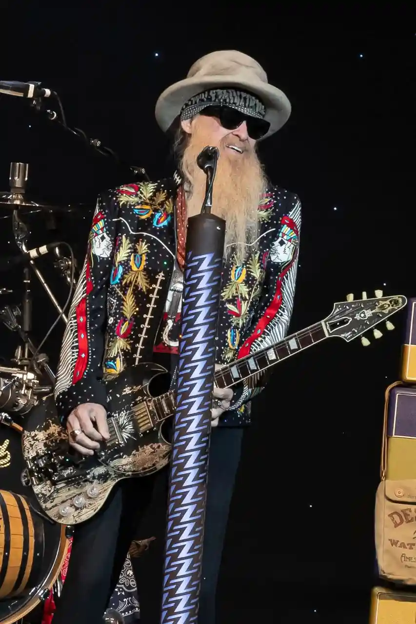 A man with a long beard, wearing a hat and sunglasses, stands on stage holding a guitar and a microphone stand covered in…